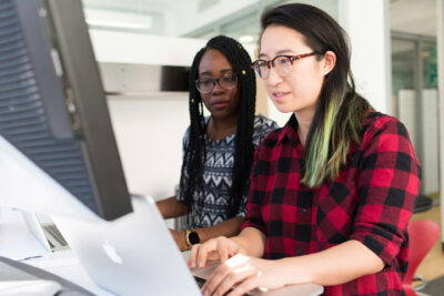Two women focused on a computer screen in an office. One wears glasses and a red plaid shirt, the other has braids and a patterned sweater.