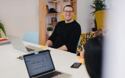 A man in glasses smiles at a desk with laptops, notebooks, and a smartphone. A relaxed, collaborative office setting with warm tones and plants.